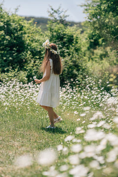 Young girl wearing daisy flower crown walks through sunny field of white blossoms during bright summer day