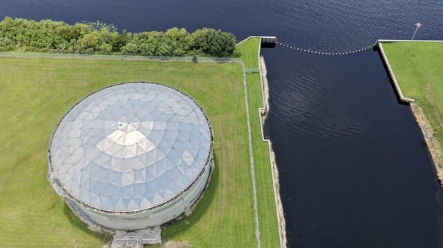 Aerial view of the FPL Sanford Power Plant featuring a large circular storage tank adjacent to a dark water channel, surrounded by green grass in DeBary, Florida, United States.