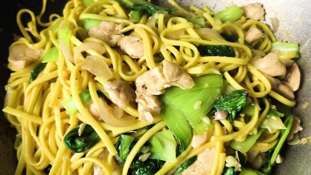 A close-up shot of steaming yellow egg noodles stir-fried to perfection with tender chicken breast pieces and green bok choy veggies.  