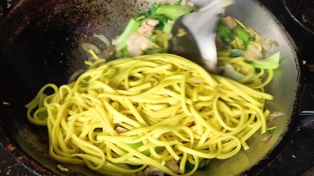 Close-up of chef hands using spatula to stir-fry yellow egg noodles with sliced chicken breast and fresh green bok choy in a traditional hot wok.  