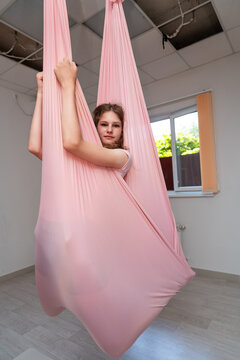 Aerial silk practice, teenage girl posing inside pink hammock during fitness session inside bright modern training studio