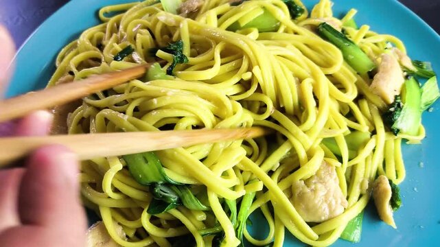 Close-up of wooden chopsticks picking up savory yellow egg noodles stir-fried with chicken and green bok choy on a blue plate.