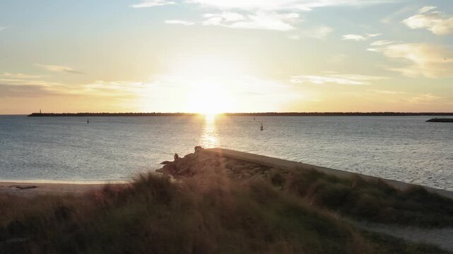 Stunning golden sunset over calm ocean waters with people fishing on concrete pier in coastal landscape.