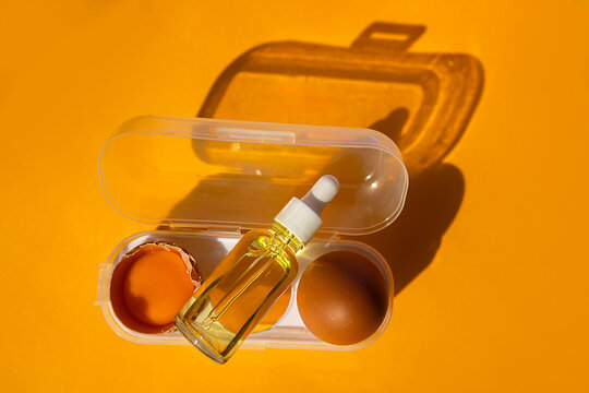 Transparent container holding a dropper bottle of oil, a cracked egg, and a whole egg on a vibrant yellow background with distinct shadows