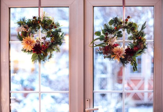 Two decorative holiday wreaths adorned with flowers and greenery hang on a window, showcasing a snowy landscape outside during winter season with soft natural light