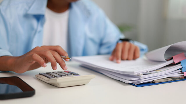 Close up male accountant hand pressing calculator button and checking financial document paperwork with stack of papers on desk. Tax invoice review and budget audit analysis concept.