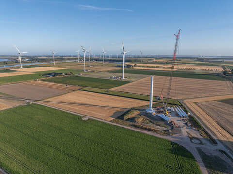 Aerial view of a wind farm construction site with a tall crane assembling a wind turbine tower amidst vast agricultural fields and existing wind turbines in The Netherlands.