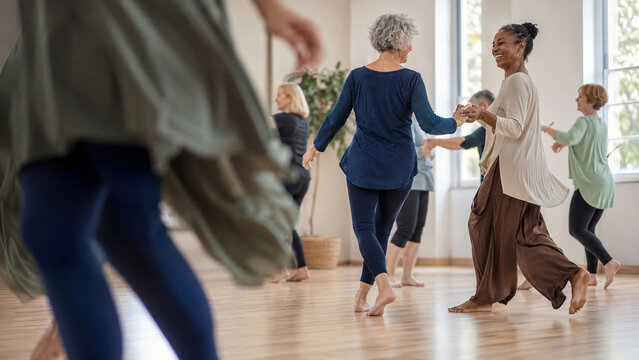 Happy senior women dance together in a bright sunlit studio, holding hands and moving barefoot across a wooden floor during a lively group exercise class with smiling active participants