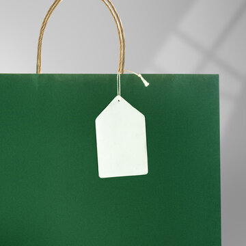 Blank white tag hanging on a dark green paper shopping bag with rope handles. Mockup or Copyspace