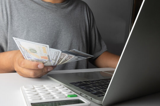 Close up of a man holding US dollar prop money while working on a laptop with a calculator. Concept of making money online, freelance income, financial planning, and home business success.
