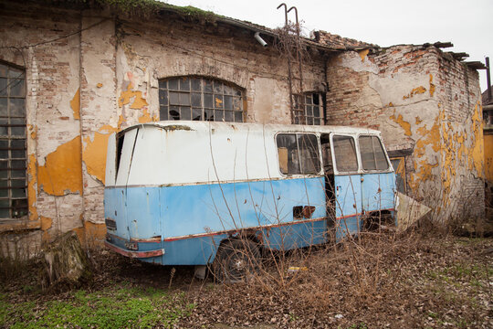 Abandoned Rusty Bus Wreck Overgrown with Weeds Beside Derelict Brick Factory Building, Urban Decay and Neglect