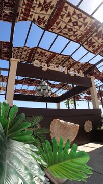 Sunlit outdoor Arabic tea and coffee kiosk near Masjid Quba with green plants.