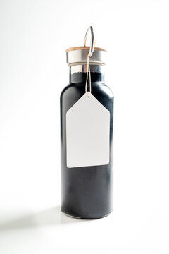 Sleek black reusable water bottle with a blank white hangtag and metal-wood cap, isolated on a white background. Mockup or Copyspace