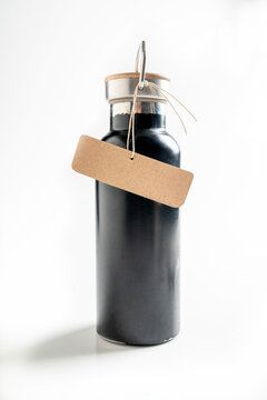 Matte black reusable stainless steel water bottle with a wooden lid and a blank kraft paper gift tag, isolated on a white background. Mockup or Copyspace