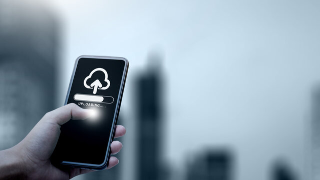 Hand holding smartphone showing cloud upload progress against blurry city background, symbolizing mobile backup and data storage. Concept of cloud computing storage and digital storage technology