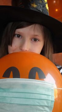 Vertical video of preteen girl in witch hat peeking from behind orange balloon with jack-o-lantern face wearing medical mask, halloween healthcare concept. Halloween peak-a-boo game