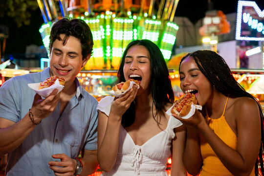 Happy young adults eating hot dogs at a night fair with bright lights