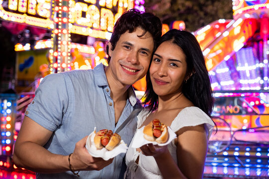 Smiling young couple holding hot dogs, enjoying an evening date at a vibrant amusement park