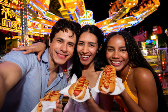 Diverse friends smiling and eating hot dogs at a vibrant amusement park fair