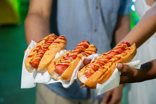 Hands holding three hot dogs with ketchup and mustard at a fair or carnival