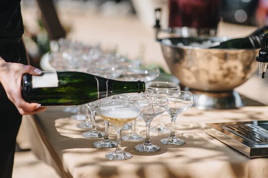 A female waitress or bartender pours bubbly champagne from a bottle into a glass on a table at a banquet. Close-up photo of food and drink. Buffet.