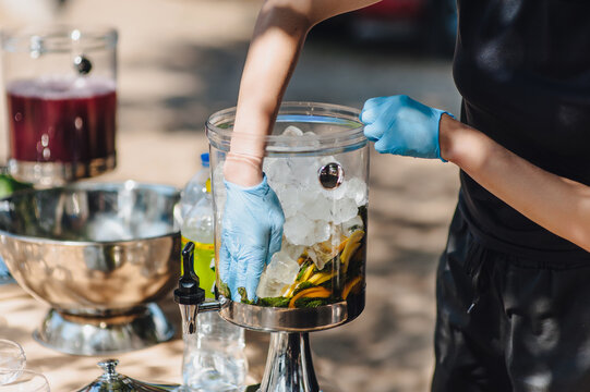 A female bartender, a waitress in gloves, prepares fresh lemonade from ice, mint, and lemon in a dispenser, a lemonade maker.