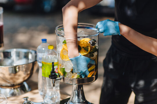 A female bartender and server wearing gloves prepare fresh lemonade from ice, mint, and lemon in a lemonade dispenser. Photo of a banquet.