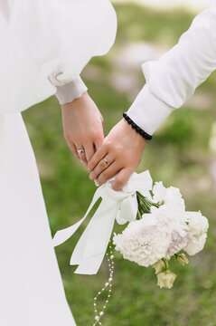 Close-up wedding photo of newlyweds, groom and bride holding hands with a bouquet of flowers outdoors, with gold rings on their fingers.