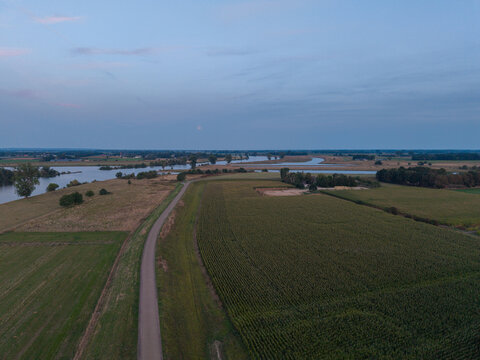 Aerial view of a winding country road cutting through vast agricultural fields and lush green meadows alongside a calm river under a soft twilight sky in The Netherlands.