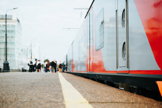 A train is positioned at a platform with visible doors and a modern design. Passengers are seen waiting on the platform under a cloudy sky, creating a busy urban atmosphere