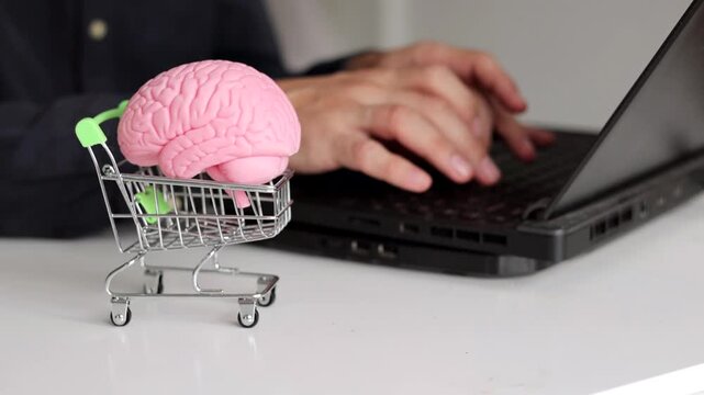 Pink brain in a miniature shopping cart beside a laptop, symbolizing online shopping, digital commerce, consumer decisions, technology and intelligent purchasing.