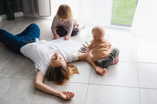 Tired mother lying on floor while her children drawing nearby.