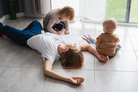 Tired mother lying on floor while her children drawing nearby.
