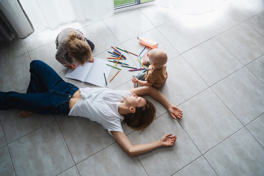 Tired mother lying on floor while her children drawing nearby.