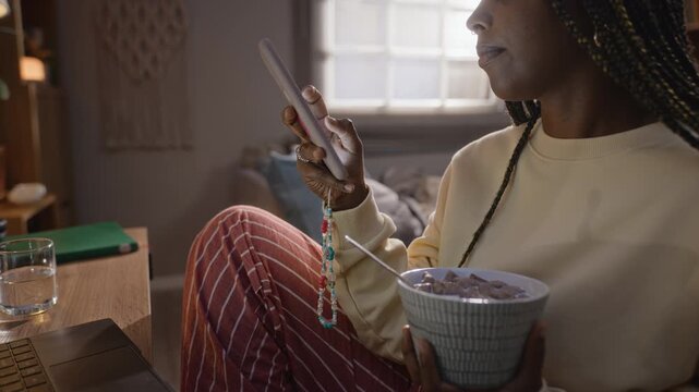 African American young adult woman holding bowl of cereal while scrolling on smartphone. Clip suitable for marketing campaigns about digital burnout, screen addiction, and remote work fatigue