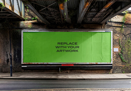 Large billboard mockup mounted on a brick wall under railway bridge in an urban street scene with industrial infrastructure, outdoor public space concrete pavement, asphalt road, railing, vines