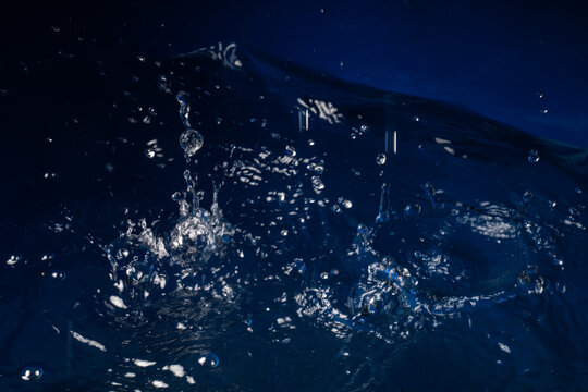 Powerful water jet splash with flying droplets on dark blue liquid background. High speed macro photo.
