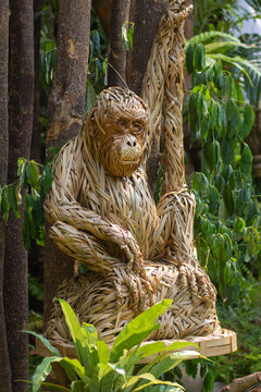 Life-sized monkey sculpture made of woven dry leaves resting among lush green tropical trees.