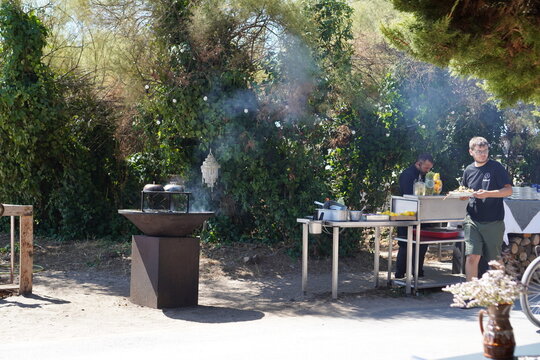A chef prepares food at an outdoor stainless steel station beside a smoking braziers. An outdoor cooking scene captures a chef at work during a catered open-air event.