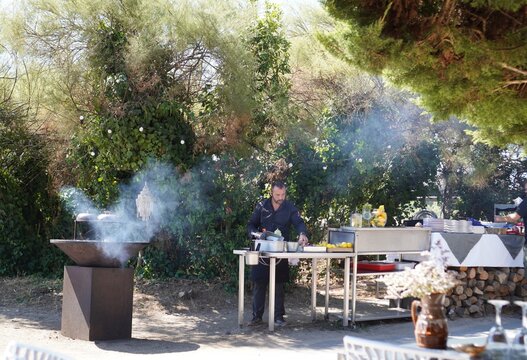 A chef prepares food at an outdoor stainless steel station beside a smoking braziers. An outdoor cooking scene captures a chef at work during a catered open-air event.