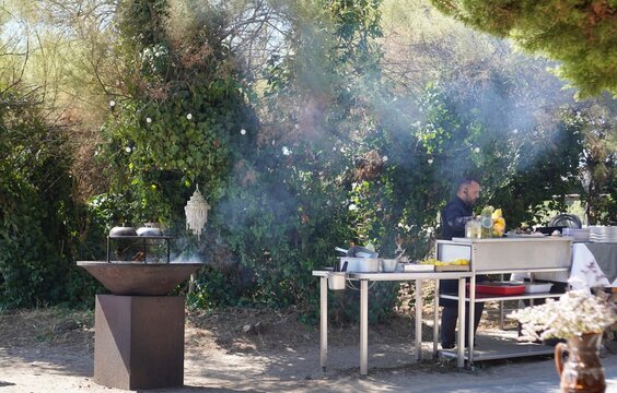 A chef prepares food at an outdoor stainless steel station beside a smoking braziers. An outdoor cooking scene captures a chef at work during a catered open-air event.