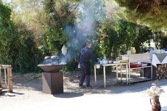 A chef prepares food at an outdoor stainless steel station beside a smoking braziers. An outdoor cooking scene captures a chef at work during a catered open-air event.