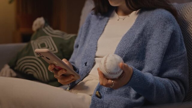 Young adult Caucasian woman sitting on sofa, holding smartphone and tossing baseball in hand. Footage is ideal for digital burnout, screen addiction, and mental wellbeing campaigns