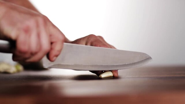 Chef hand crushing fresh garlic clove with steel knife blade on wooden kitchen surface.