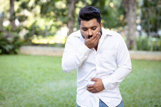 young adult south asian man in his 20s feeling nauseous and about to vomit in outdoors park, holding his stomach in distress