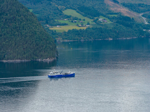 Aerial view of a blue wellboat navigating the calm waters of a fjord surrounded by lush green hills in Stranda, More og Romsdal, Norway.