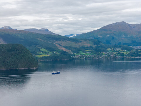 Aerial view of a wellboat navigating the calm waters of a deep fjord surrounded by lush green mountains and rolling hills in Stranda, More og Romsdal, Norway.