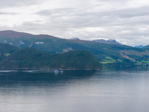 Aerial view of a wellboat navigating the calm waters of a fjord, surrounded by lush green mountains and rolling hills under a cloudy sky in Stranda, More og Romsdal, Norway.