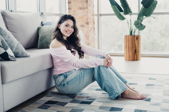 Smiling young woman sitting on the floor in a cozy modern living room wearing a pink off-shoulder sweater and jeans, relaxed and comfortable at home