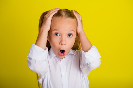 Surprised little girl with hands on head showing shock and astonishment in white shirt and ponytails against a bright yellow background
